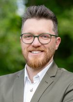 Porträt einer lächelnden Person mit Brille, Bart und Sakko vor grünem Hintergrund.
